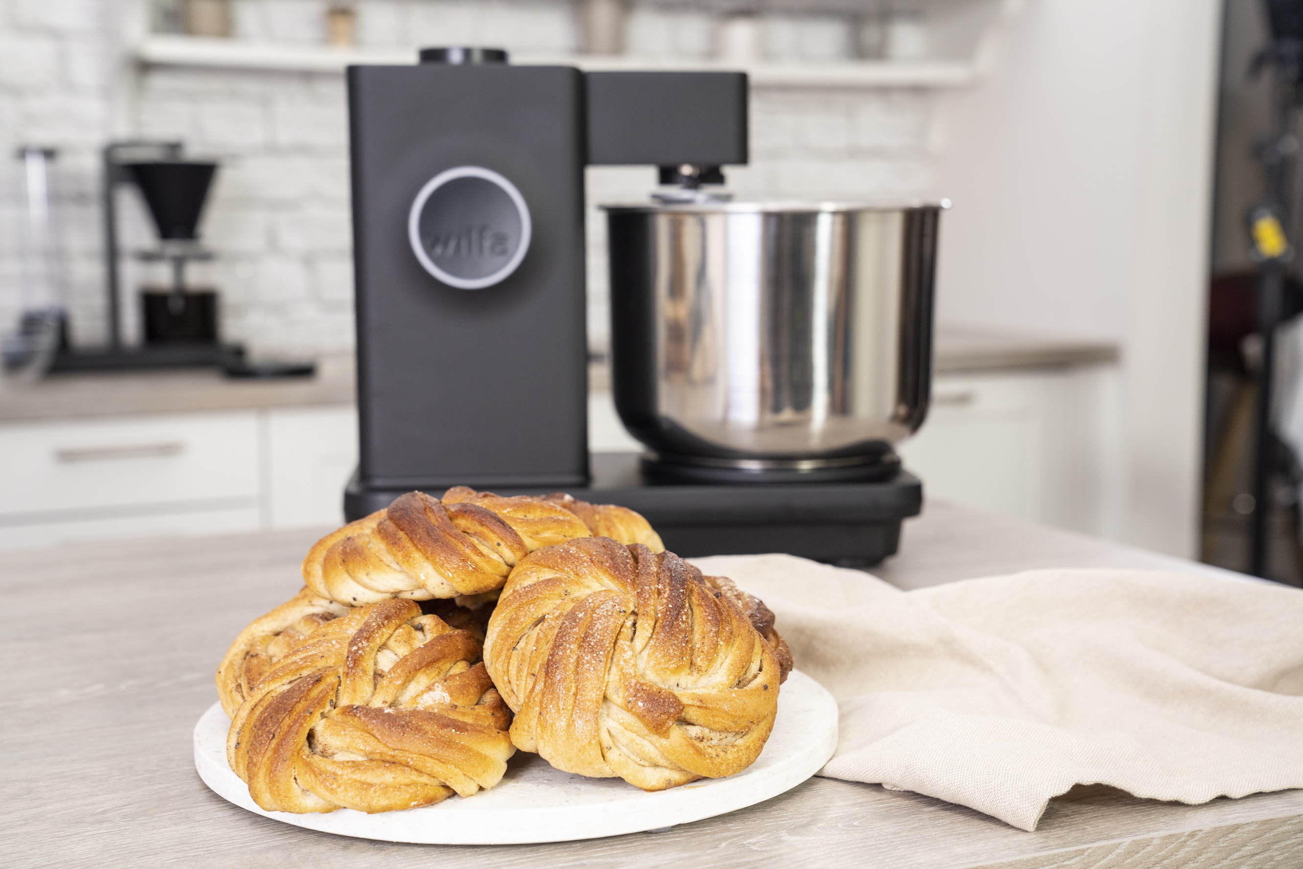 Küchenmaschine mit Edelstahlbehälter, im Hintergrund knusprige Zimtschnecken auf einem Teller, schöne, moderne Kücheneinrichtung.
