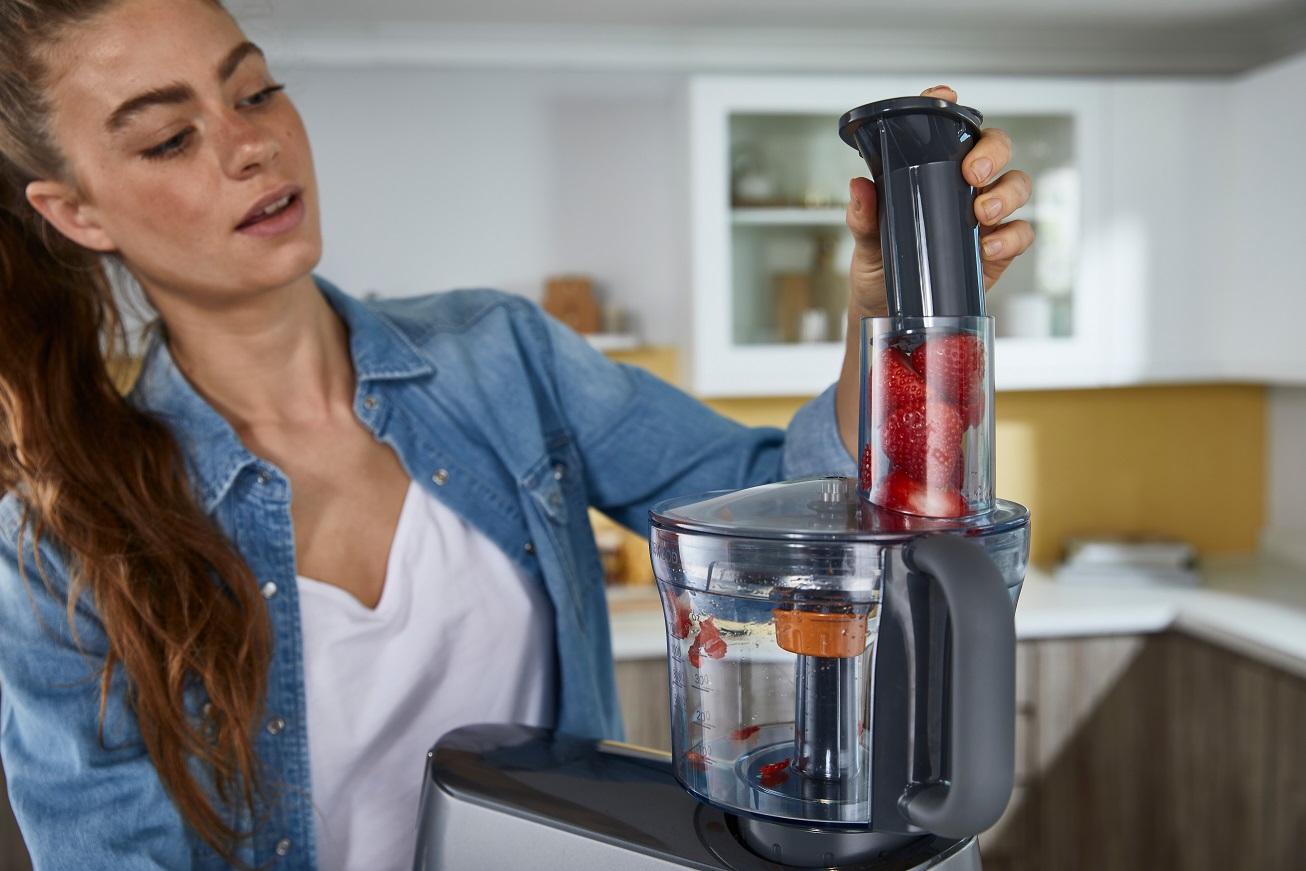 Frau mit Küchenmaschine, füllt sie mit frischen Erdbeeren zur Zubereitung. Modernes Küchendesign im Hintergrund.